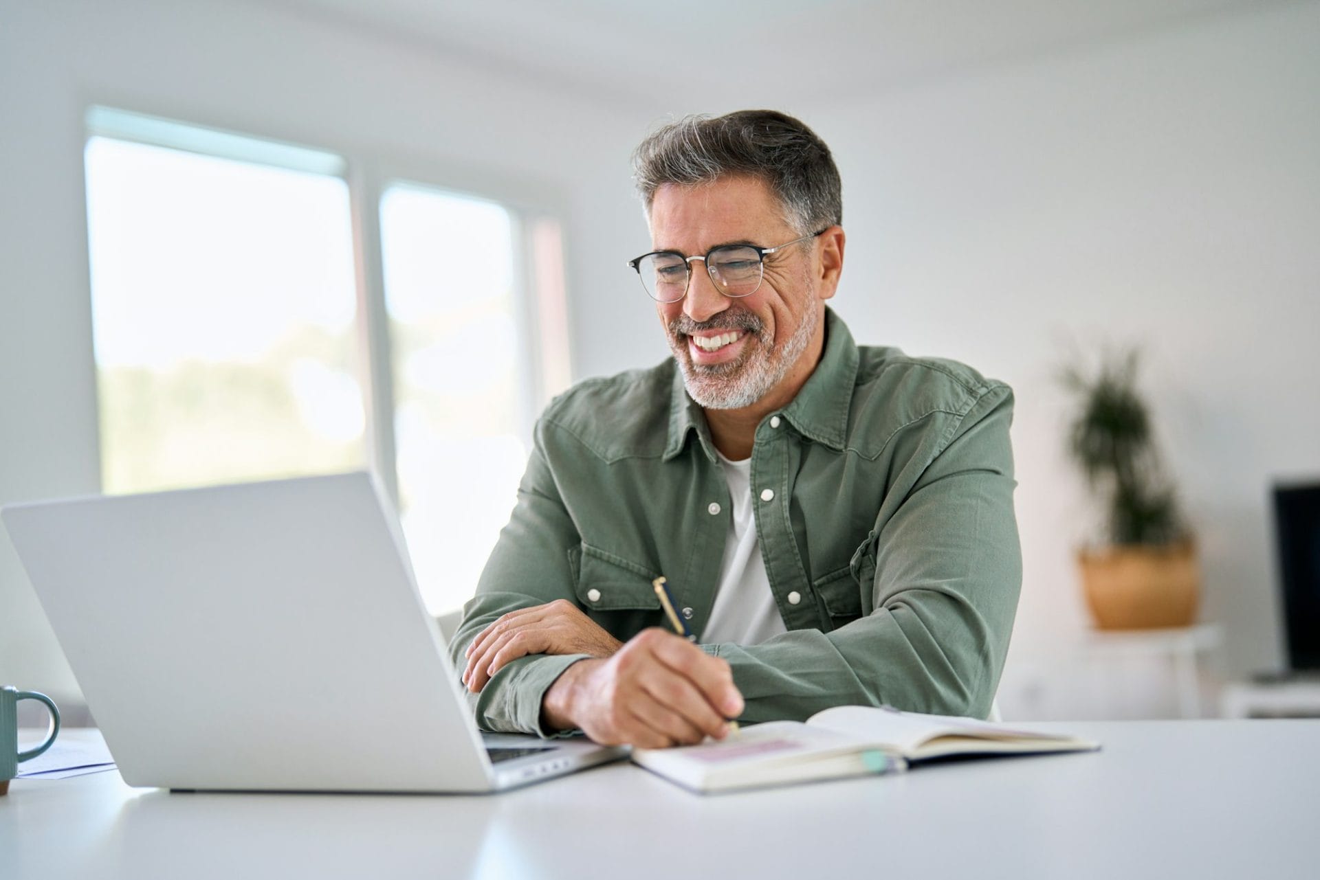 Happy middle aged man using laptop computer writing notes sitting at home table.